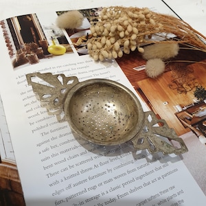 May include: Antique silver tea strainer with a perforated bowl and ornate leaf-shaped handles. The strainer sits on a magazine page with text and dried flowers. A traditional kitchen accessory.