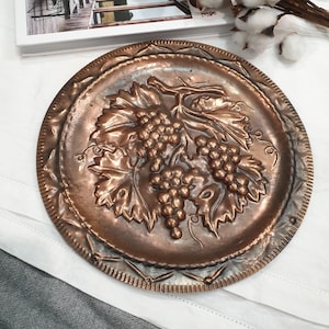 May include: A circular, copper-toned decorative plate featuring a raised relief of grapes and leaves. The plate has a detailed border and a vintage appearance. The grapes and leaves are the central design element, creating a classic, rustic aesthetic.