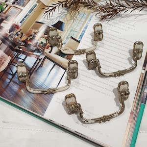 May include: A set of antique cabinet pulls with a distressed white finish and ornate, aged metal details. These vintage drawer handles feature a curved design, perfect for furniture restoration projects.