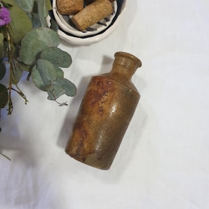May include: A rustic brown ceramic bottle with a wide mouth and a slightly rounded shape. The bottle has a rough, textured surface and appears to be vintage.