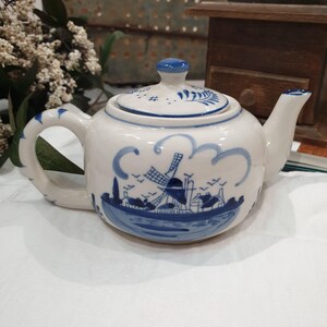 May include: A white ceramic teapot featuring a blue hand-painted landscape design of a windmill, houses, and birds. The teapot has a matching lid with a blue knob, a curved handle, and a spout, all decorated with blue floral and line accents.