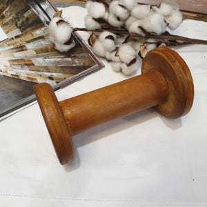 May include: A wooden spool with a cylindrical body and two circular ends. The spool is a warm brown colour and appears to be made of solid wood. The spool is lying on a white surface, with cotton bolls and a magazine in the background.