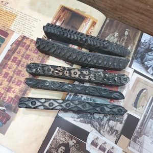 May include: Five dark, carved wooden decorative pieces. Each piece has a unique pattern, including floral and geometric designs. The pieces are arranged on a vintage book with old photographs.