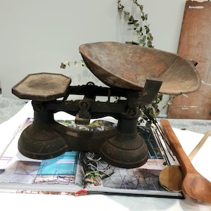 May include: A vintage cast iron scale, featuring a large, curved tray and a smaller, octagonal platform. The scale has a dark brown, weathered finish. Two wooden spoons are placed nearby. The scale's design suggests it is for measuring ingredients in a kitchen.