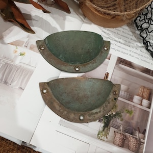 May include: Two semi-circular metal drawer pulls with a weathered, oxidised green patina. Each pull features a recessed cup design and three pre-drilled screw holes along the flat edge for mounting, showing signs of age and surface wear.