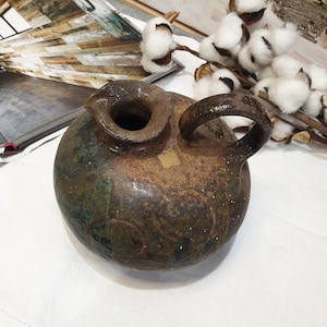 May include: A handcrafted, rustic ceramic jug with a rounded body and a handle. The jug features a mottled brown and green glaze with a textured surface. The opening is circular, and the overall design suggests a vintage or antique aesthetic.