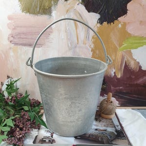 May include: A vintage galvanized metal bucket with a curved handle. The bucket has a cylindrical shape with three horizontal lines. The background features an abstract painting with shades of pink, brown, and yellow. A bundle of purple flowers and green leaves are on the left.