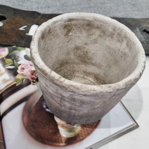 May include: A grey concrete planter with a textured finish. The open top reveals the interior, and the planter sits on a magazine with floral imagery. The planter's design suggests a rustic or minimalist aesthetic.
