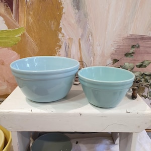 May include: Two light blue ceramic mixing bowls with a smooth, glossy finish and a rounded shape. The larger bowl is positioned to the left, while the smaller bowl sits to the right. Both bowls feature a simple, clean design with a subtle rim detail.