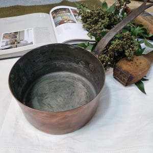 May include: A vintage copper saucepan with a long, metal handle. The pan has a rounded shape and is on a white surface. The interior shows signs of use. A wooden stamp and open book are in the background.