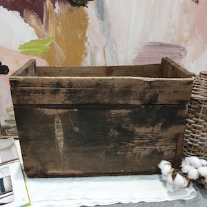 May include: A rustic, rectangular wooden box with a dark brown finish. The box has a simple, open design with visible wood grain and signs of wear. The background features an abstract painting with pink, brown, and green hues. A woven basket and cotton bolls are also visible.