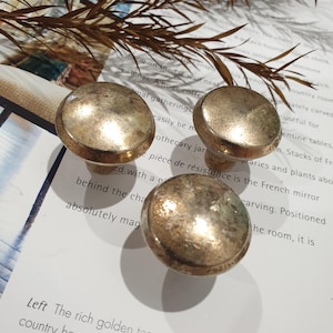 May include: Four antique-style cabinet knobs with a tarnished gold finish. The knobs are round with a slightly domed top and a short, cylindrical base. They are arranged on a white surface with text and a sprig of dried foliage.