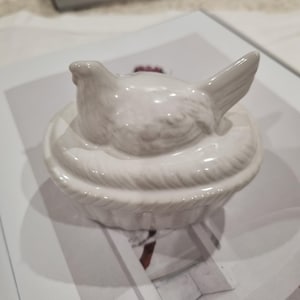 May include: A white ceramic lidded dish featuring a sculpted bird sitting atop a textured nest-like base. The glossy finish covers the entire piece, which includes a detailed bird figure with wings and a tail, resting on a woven-patterned container.