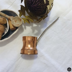 May include: A small copper coffee pot with a brass handle. The pot is sitting on a white surface.