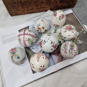 May include: Eight white ceramic spheres with floral designs in various colours. Some have small holes and decorative elements. The spheres are arranged on a magazine page, showcasing a vintage aesthetic.