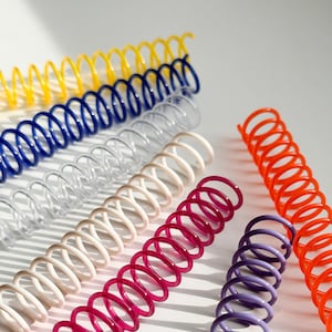 May include: Assortment of colorful plastic binding coils in yellow, blue, clear, white, pink, purple, and orange. The coils are arranged in a row on a white surface.