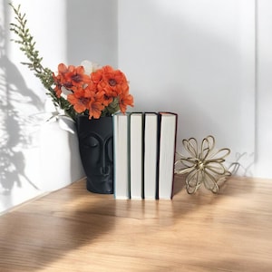 May include: A black ceramic vase with a face design, filled with orange and white flowers, sits on a wooden table next to a stack of five books and a gold metal decorative flower.