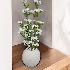 Puede incluir: Jarrón de cerámica blanco con una superficie texturizada en forma de esfera. El jarrón está lleno de un ramo de flores blancas con tallos verdes.