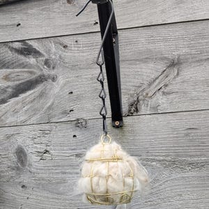 May include: A black metal hanging bird feeder with a gold wire cage filled with light-colored nesting material. The feeder is attached to a wooden wall with visible wood grain.