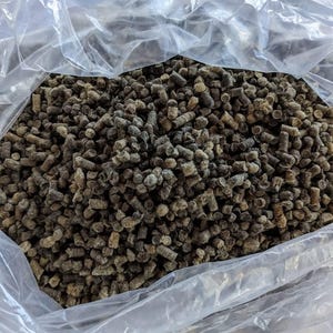 May include: A transparent plastic bag overflowing with numerous small, cylindrical, brown pellets. The pellets are textured and display varying shades of brown and grey. The bag is partially open, exposing the contents.