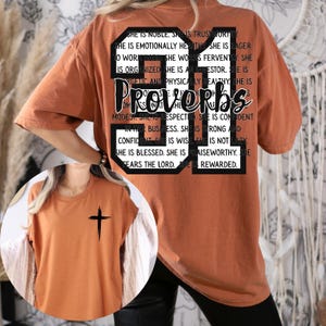 May include: Burnt orange t-shirt with a black cross graphic on the front. The back of the shirt features the text "Proverbs 31" in a black box, with additional text describing the qualities of a virtuous woman.
