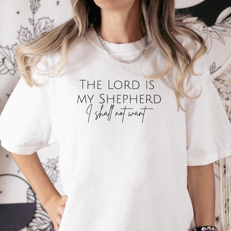 Puede incluir: Camiseta blanca con el texto "THE LORD IS MY SHEPHERD I shall not want" en escritura negra. La camiseta tiene cuello redondo y mangas cortas. La modelo lleva un collar plateado.
