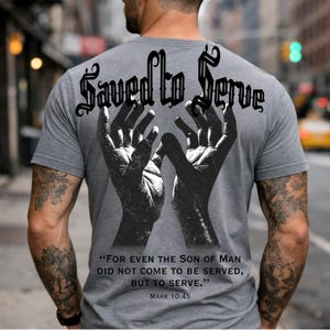 May include: Gray t-shirt with the words "Saved to Serve" in a gothic font and a black and white graphic of two hands reaching upwards. The shirt also features a quote from Mark 10:45.