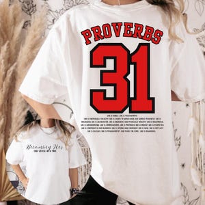 May include: White t-shirt with the text "PROVERBS 31" in red and black. The number 31 is large and prominent. Below the number is a list of positive attributes. A second shirt with the text "Becoming Her" is also visible.