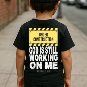May include: Black t-shirt with a yellow and black construction sign graphic. The sign reads "UNDER CONSTRUCTION" above "GOD IS STILL WORKING ON ME" in white text. The Bible verse "EPHESIANS 2:10" is below the text.