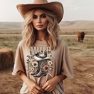 May include: A tan t-shirt with a graphic of a cowboy boot and hat with flowers. The text "Wild and Free" is printed on the shirt.