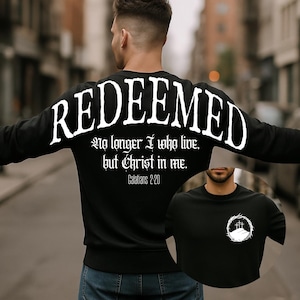 May include: Black long-sleeve sweatshirt with the word "REDEEMED" in large white letters across the back. Below the word is the text "No longer I who live, but Christ in me. Galatians 2:20". A smaller image of a black t-shirt is visible.