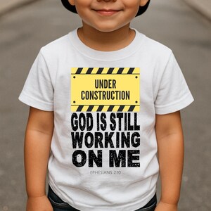 May include: White t-shirt with a yellow and black "Under Construction" sign graphic. Below the sign, the shirt displays the text "GOD IS STILL WORKING ON ME" in black, with "Ephesians 2:10" below. The shirt is for children.