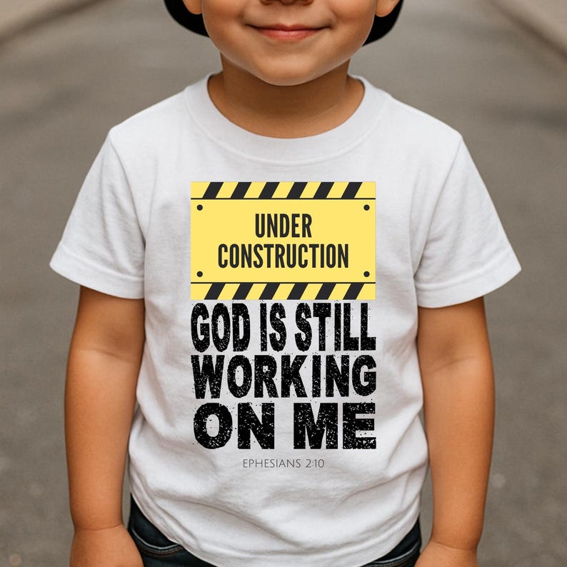 May include: White t-shirt with a yellow and black "Under Construction" sign graphic. Below the sign, the shirt displays the text "GOD IS STILL WORKING ON ME" in black, with "Ephesians 2:10" below. The shirt is for children.