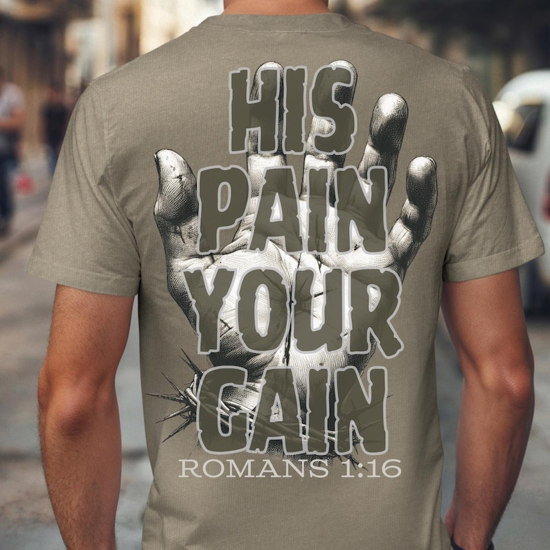 Puede incluir: Camiseta color arena con un gr&aacute;fico en la espalda que presenta una mano detallada con una corona de espinas y las palabras "HIS PAIN YOUR GAIN" en una fuente desgastada. El texto "ROMANS 1:16" est&aacute; debajo.
