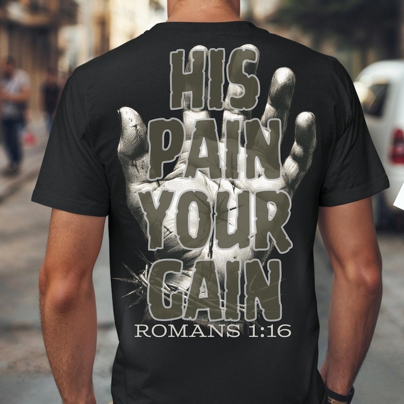 Puede incluir: Camiseta negra con un dise&ntilde;o gr&aacute;fico en la espalda. El dise&ntilde;o presenta una imagen detallada en escala de grises de una mano con las palabras "HIS PAIN YOUR GAIN" en una fuente desgastada. El texto "ROMANS 1:16" est&aacute; debajo.