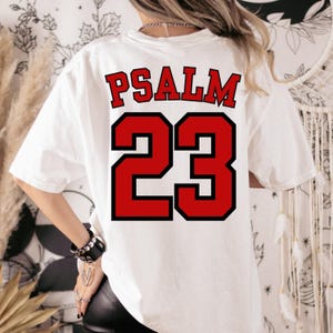 Puede incluir: Camiseta blanca con el texto "PSALM 23" en letras rojas con contorno negro. El n&uacute;mero "23" tambi&eacute;n es rojo con contorno negro. La camiseta es de estilo casual.