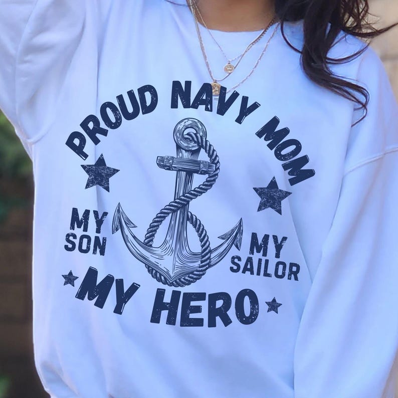 May include: White sweatshirt with a navy blue anchor design and the text "PROUD NAVY MOM" in a circular arrangement. The design also includes the phrases "MY SON", "MY SAILOR", and "MY HERO", along with star accents.