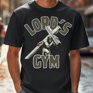Puede incluir: Camiseta negra con un dise&ntilde;o gr&aacute;fico que presenta una figura musculosa que lleva una cruz. El texto "LORD'S GYM" est&aacute; encima y debajo de la imagen, con "THE SIN OF THE WORLD" en la cruz. El dise&ntilde;o es en tonos grises y blancos.