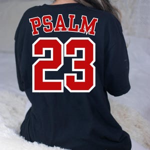 Puede incluir: Una camiseta negra con el texto "PSALM 23" en letras rojas con contorno blanco. La camiseta se ve desde la espalda, con el texto centrado en la parte superior de la espalda.