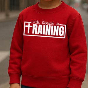 Könnte beinhalten: Ein rotes Sweatshirt mit dem weißen Text "Little Disciple in TRAINING" in einem weißen, rechteckigen Rahmen. Das Sweatshirt hat eine einfarbige rote Farbe und scheint aus einem weichen Material zu sein. Der Text ist fett und ohne Serifen.