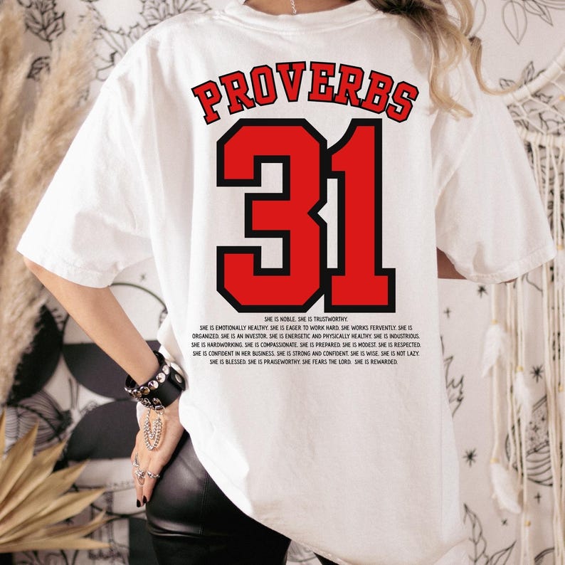 May include: White t-shirt with the text "PROVERBS 31" in red and black. The number 31 is large and red. Below the number is a list of qualities. A casual, comfortable tee.