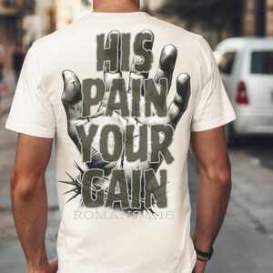 Puede incluir: Una camiseta color crema con un dise&ntilde;o gr&aacute;fico en la espalda. El dise&ntilde;o presenta una ilustraci&oacute;n detallada de una mano con las palabras "HIS PAIN YOUR GAIN" en una fuente desgastada. El texto "ROMANS 12:16" est&aacute; debajo.