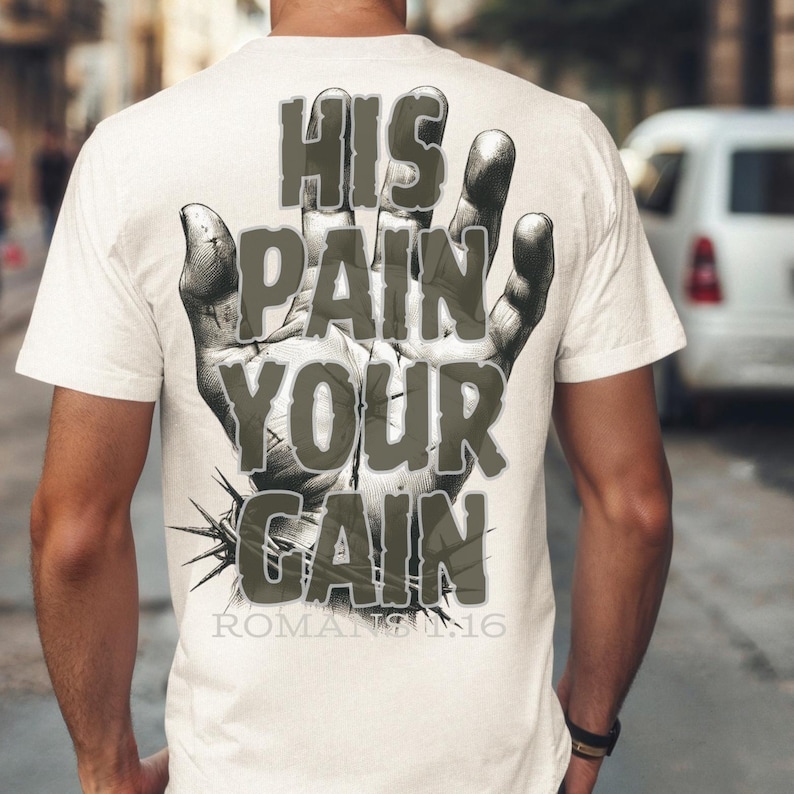 Puede incluir: Una camiseta color crema con un dise&ntilde;o gr&aacute;fico en la espalda. El dise&ntilde;o presenta una ilustraci&oacute;n detallada de una mano con las palabras "HIS PAIN YOUR GAIN" en una fuente desgastada. El texto "ROMANS 12:16" est&aacute; debajo.