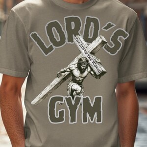 Puede incluir: Camiseta color beige con la frase "LORD'S GYM". El dise&ntilde;o muestra una figura musculosa cargando una cruz, con el texto "The Sin of the World" en la cruz.