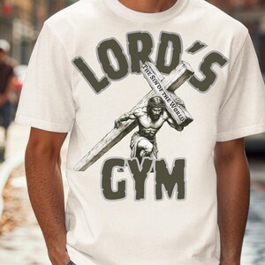 Puede incluir: Camiseta blanca con un dise&ntilde;o gr&aacute;fico con el texto "LORD'S GYM". El dise&ntilde;o incluye una ilustraci&oacute;n de un hombre cargando una cruz, con las palabras "THE SIN OF THE WORLD" en la cruz.