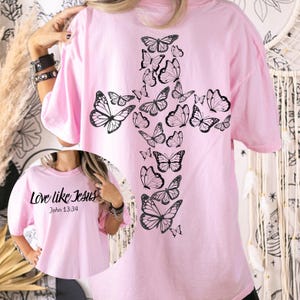 May include: A pale pink t-shirt with a cross design made of black butterflies on the back. The front displays the text "Love like Jesus" and "John 13:34". The shirt has short sleeves and a loose fit.