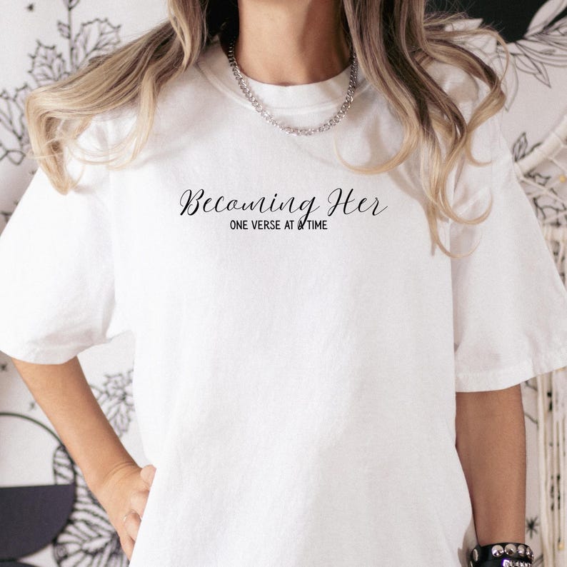May include: White t-shirt with the text "Becoming Her" in a cursive font above the text "ONE VERSE AT A TIME" in a smaller font. The shirt has short sleeves and a crew neck.
