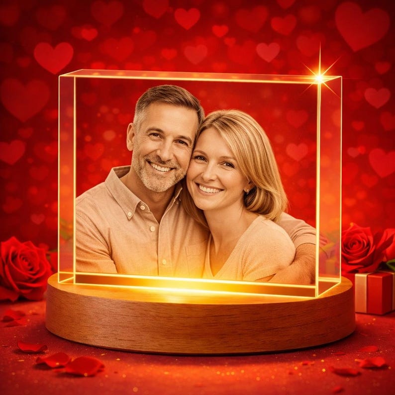 Puede incluir: Un bloque de vidrio rectangular con un retrato grabado con l&aacute;ser de una pareja sonriente, iluminado por una fuente de luz c&aacute;lida. El bloque de vidrio est&aacute; montado sobre una base circular de madera, sobre un fondo rojo vibrante con corazones.