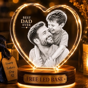 May include: A heart-shaped clear acrylic plaque featuring a black and white photo of a father and son, with the text BEST DAD EVER. The plaque sits on a round wooden base with a metal plate reading FREE LED BASE, illuminated by warm light.