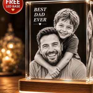 Puede incluir: Un bloque de vidrio rectangular con una fotograf&iacute;a grabada con l&aacute;ser en blanco y negro de un adulto y un ni&ntilde;o sonrientes abrazados. El texto BEST DAD EVER est&aacute; grabado sobre las figuras. El vidrio descansa sobre una base de madera LED.